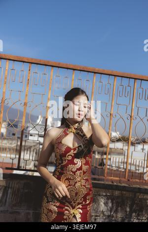 Gracieuse jeune femme asiatique dans la robe rouge cheongsam se sentant sereine et paisible sur le toit. modèle élégant bénéficie de la lumière du soleil sous un ciel bleu clair dans un urb Banque D'Images