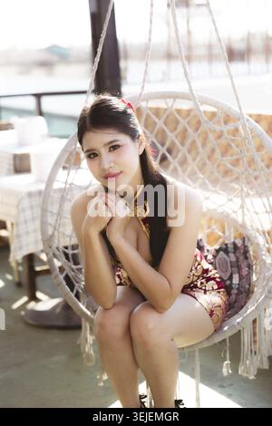 Élégante jeune femme chinoise en robe qipao moderne se trouve dans une chaise pivotante suspendue à l'extérieur. Elle a un look calme et serein avec une expression douce et invitante Banque D'Images