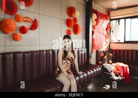 Femme asiatique élégante dans la robe rouge Cheongsam assise sur le canapé en cuir. Beau modèle féminin posant avec une expression calme lors de la célébration du nouvel an chinois Banque D'Images