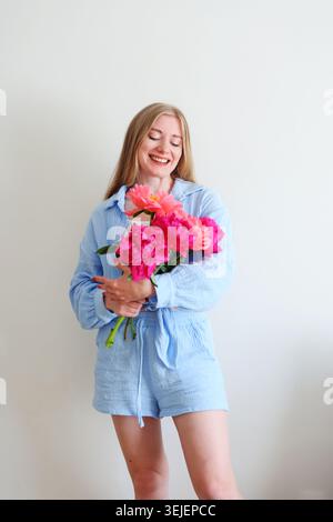 Jeune femme souriante tenant des pivoines roses vibrantes dans un vase en verre, portrait de style de vie doux avec une humeur printanière fraîche et un fond minimaliste. Banque D'Images