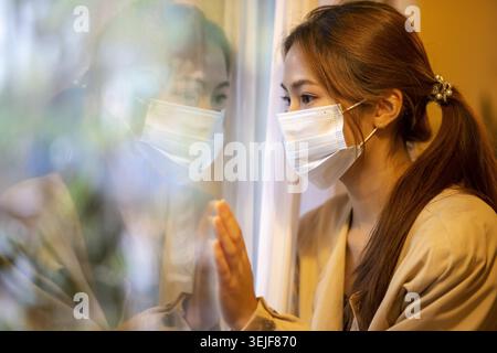 Femme asiatique triste et solitaire portant un masque en quarantaine à la maison, regardant par la fenêtre avec son reflet. sentiment d'isolement au cours d'une pandémie mondiale Banque D'Images