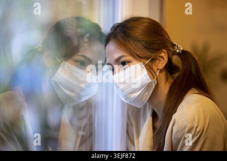 Triste jeune femme asiatique dans le masque facial se sentant seule pendant la quarantaine. portrait d'isolement et de dépression, elle regarde par la fenêtre avec reflec réfléchi Banque D'Images