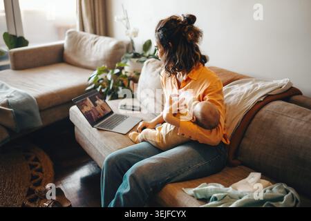 Une mère en chemise orange est assise sur un canapé et nourrit son bébé avec un biberon tandis qu'un ordinateur portable montre un appel vidéo, capturant un moment agréable à la maison. Banque D'Images