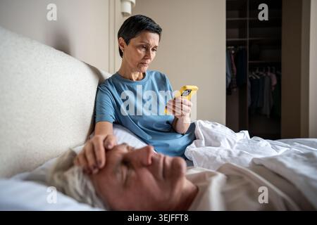 Femme âgée inquiète à la recherche de signes de maladie par téléphone sentant sourcil de mari malade se reposant dans le lit Banque D'Images
