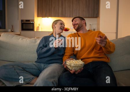 Rire couple heureux manger du pop-corn et rire tout en regardant la télévision ensemble à la maison sur le canapé Banque D'Images