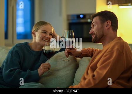 Couple heureux appréciant le vin ensemble dans le salon confortable partageant une conversation intime sur le canapé la nuit Banque D'Images
