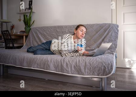 Détendu shopping en ligne de la femme tout en étant à l'aise couché avec la carte bancaire sur le canapé, en utilisant l'ordinateur portable. Banque D'Images