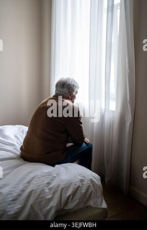 Homme âgé assis seul dans le fardeau émotionnel fatigue mentale et dépression profonde par fenêtre, rétrovision Banque D'Images