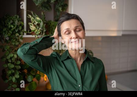 Femme mature satisfaite appréciant la coupe de cheveux courte à la maison en courant la main à travers les cheveux, senior slow Life Banque D'Images