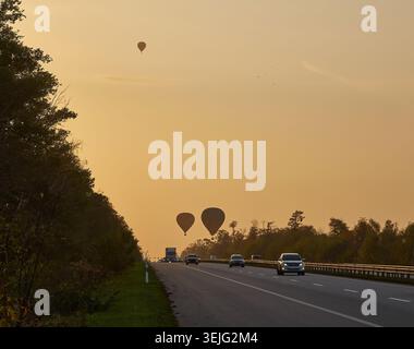 Deux montgolfières silhouettées contre un ciel brumeux de lever ou coucher de soleil, flottant au-dessus d'une autoroute achalandée avec des voitures et des camions. Des arbres bordent le bord de la route. Banque D'Images