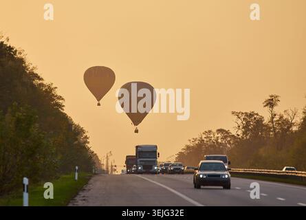 Deux montgolfières silhouettées contre un ciel brumeux de lever ou coucher de soleil, flottant au-dessus d'une autoroute achalandée avec des voitures et des camions. Des arbres bordent le bord de la route. Banque D'Images