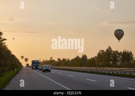 Deux montgolfières silhouettées contre un ciel brumeux de lever ou coucher de soleil, flottant au-dessus d'une autoroute achalandée avec des voitures et des camions. Des arbres bordent le bord de la route. Banque D'Images