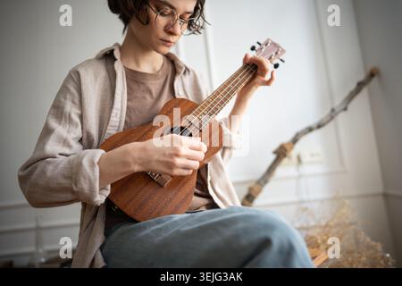 Jeune femme apprenant un nouveau passe-temps, jouant sur ukulélé en bois, plumant des cordes, aime le processus créatif. Banque D'Images