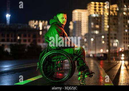 Homme handicapé en fauteuil roulant dans la lumière rgb néon vert posant sur fond de ville illuminé Banque D'Images