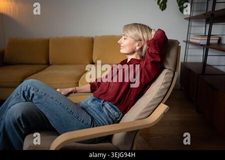 Heureuse satisfaite femme d'âge moyen se reposant, rêvant dans une chaise confortable à la maison, profitant pleinement de la détente Banque D'Images