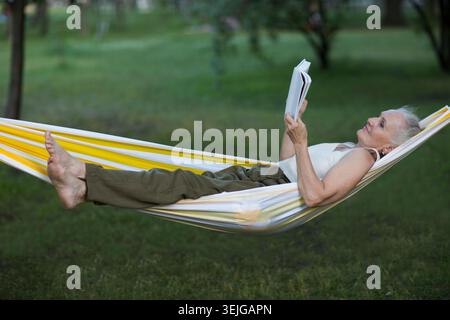Femme âgée avec les cheveux gris se repose et lit un livre couché dans un hamac Banque D'Images