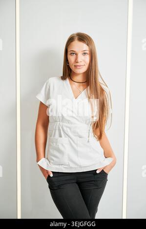 Femme souriante dentiste se tient dans un bureau moderne portant un haut de gommage blanc. Portrait médical professionnel pour la publicité clinique, les sites Web de dentisterie, la communication sur les soins de santé, la présentation du personnel Banque D'Images