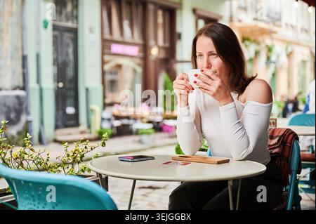 Femme sirotant un café en plein air dans la rue de la ville, assise à table avec smartphone, atmosphère urbaine confortable avec de vieux bâtiments, concept de loisirs, de voyage et de style de vie détendu Banque D'Images