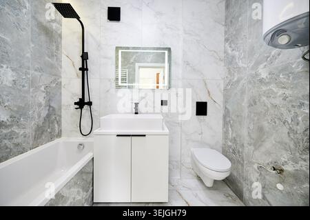 Meuble-lavabo avec lavabo, baignoire et toilettes murales dans un intérieur moderne. Les carreaux de marbre et le système de douche noir créent un espace minimal propre, un concept de rénovation et un design de maison contemporain Banque D'Images