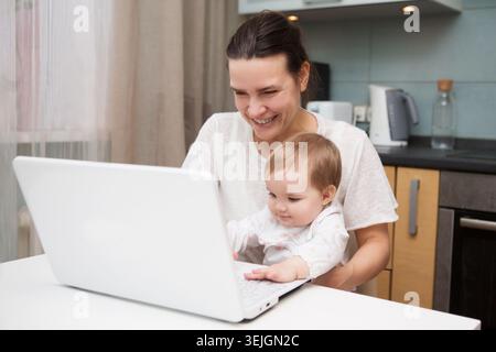 Maman et bébé. une jeune mère travaillant avec un ordinateur portable dans la cuisine Banque D'Images