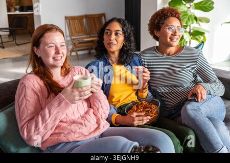 Amies féminines diverses se relaxant sur le canapé en sirotant une tasse grignotant sur des bretzels et en appuyant sur la télécommande. Confortable, décontracté, amitié, lei Banque D'Images