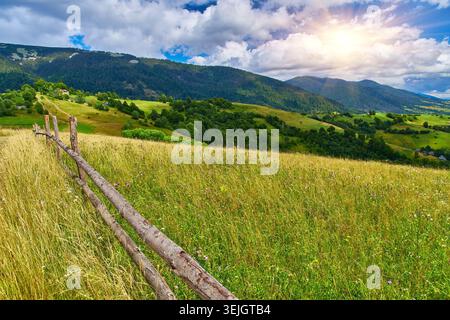 Une clôture en bois rustique traverse une pente de prairie de montagne herbeuse surplombant des collines boisées sous un ciel bleu avec des nuages spectaculaires. Terres d'été rurales Banque D'Images