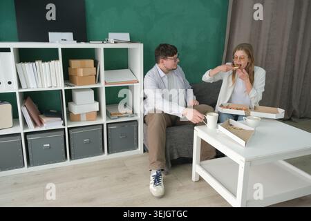 Homme et femme milléniaux travaillant dans un petit bureau, ayant une pause déjeuner avec café et pizza Banque D'Images