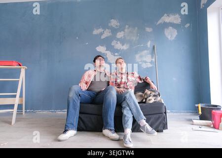 Heureux couple rénover un appartement de maison neuve, assis sur le canapé, discuter des plans ensemble et caresser un chien mignon Banque D'Images