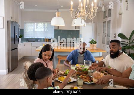 Famille diversifiée partageant des assiettes de poulet rôti autour d'une table en bois dans une cuisine moderne. Famille, banquet, salle à manger, célébration, confortable, Banque D'Images