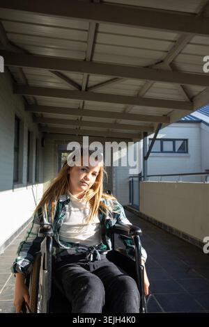 Enfant de sexe féminin assis dans un fauteuil roulant manuel naviguant dans un couloir carrelé sous des poutres de toit en bois. Accessibilité, enfance, extérieur, Banque D'Images