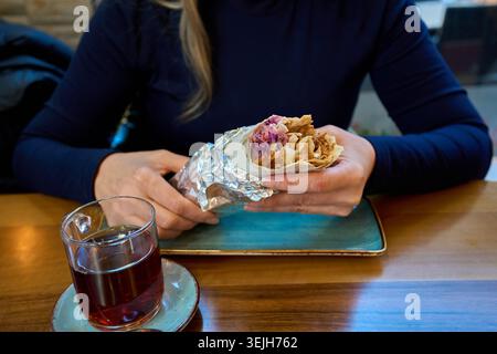 Une femme tenant un wrap shawarma au poulet en papier aluminium sur une table en bois avec une tasse en verre de thé noir dans un café. Banque D'Images