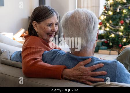 Couple senior assis étroitement ensemble sur le canapé dans le salon avec cheminée et arbre de Noël. Confortable, chaleur, intimité, celea Banque D'Images