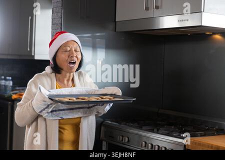 Femme asiatique senior portant chapeau de Père Noël et tenant un plateau de cuisson avec des biscuits dans la cuisine, espace de copie. Cuisson maison, saison des fêtes, f Banque D'Images