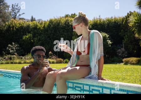 Couple diversifié portant des lunettes de soleil assis sur le bord de la piscine carrelée et debout dans l'eau retenant des boissons. Luxe, détente, loisirs, Banque D'Images