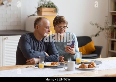 Couple senior prenant le petit déjeuner et utilisant un smartphone à la maison Banque D'Images