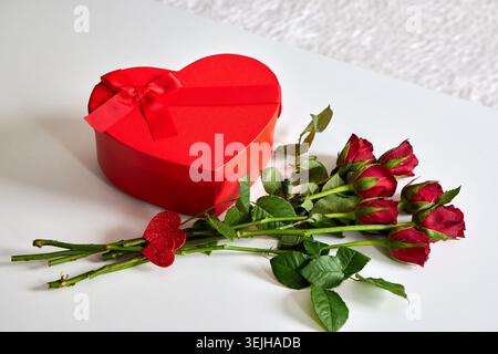 Une boîte cadeau festive en forme de cœur rouge avec un noeud en ruban couché à côté d'un bouquet de roses rouges fraîches. Banque D'Images