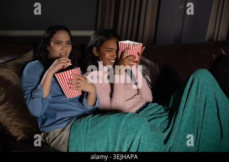 Diverses amies féminines regardant un film de suspense sur un canapé sous une couverture verte tenant des seaux de pop-corn. Confort, amitié, détente Banque D'Images