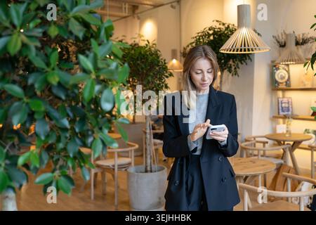 Femme d'affaires positive qui a réussi le directeur de restaurant envoyant des SMS sur son smartphone pendant la pause de travail. Banque D'Images