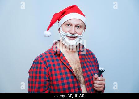 Un homme souriant dans un chapeau de Père Noël et une chemise à carreaux avec de la mousse à raser sur son visage tenant un rasoir. Banque D'Images