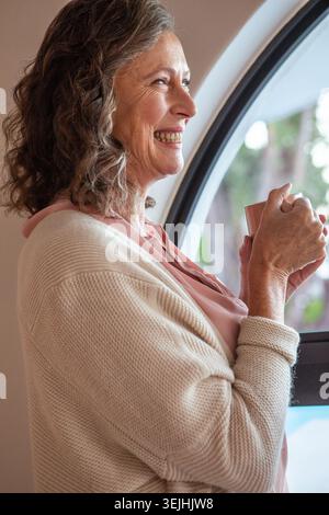 Femme senior debout près de la fenêtre cintrée à l'intérieur de la maison tenant tasse portant cardigan, espace de copie. Confiant, confortable, sérénité, domest Banque D'Images