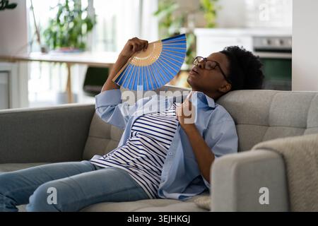 Une femme afro-américaine fatiguée et épuisée s'assoit sur un canapé en utilisant des fans de papier souffrant de stuffess Banque D'Images