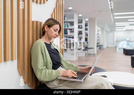 Femme ciblée auteur indépendant travaillant sur ordinateur portable dans la bibliothèque. Pigiste après 40 Banque D'Images