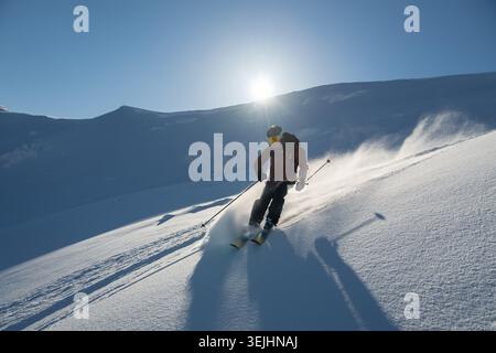 Un skieur sculpte à travers la poudre fraîche sur une pente ensoleillée, entourée par le ciel bleu et les montagnes. Le groupe aime l'aventure hors-piste avec un hélicoptère-Li Banque D'Images