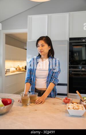 Femme asiatique tenant deux verres de jus tout en se tenant debout à l'îlot de cuisine avec des légumes frais. Moderne, minimaliste, vibrant, nous Banque D'Images