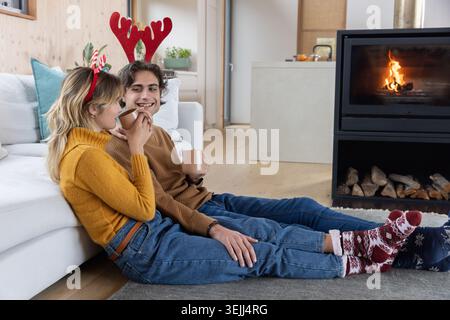 Couple assis sur un tapis dans le salon près de la cheminée portant des bandeaux en bois tenant des tasses en céramique. Confort, intimité, célébrati Banque D'Images