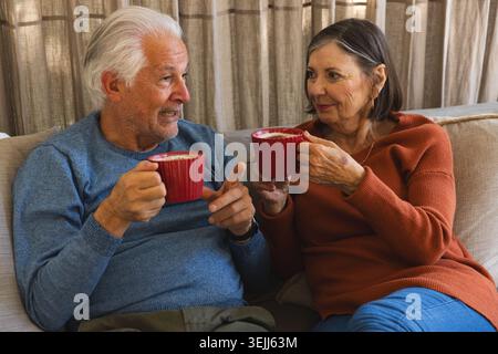 Un couple âgé est assis sur le canapé dans le salon, buvant des tasses en céramique rouge et bavardant. Cosiness, convivialité, leis Banque D'Images