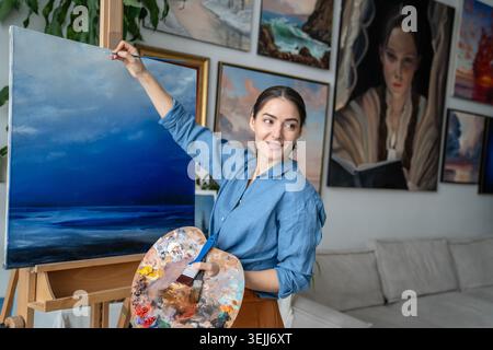 Femme talentueuse souriante artiste peignant image sur toile à l'atelier dans une atmosphère détendue positive. Banque D'Images