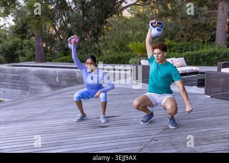 Divers partenaires d'entraînement assis en position squat sur une terrasse en bois à côté de la piscine avec kettlebells au-dessus de leurs têtes. F Banque D'Images