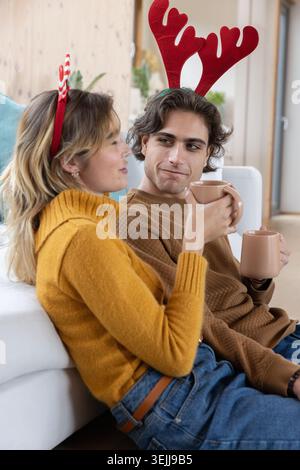 Couple assis sur le canapé dans le salon portant des bandeaux festifs en bois tenant des tasses en céramique. Confort, célébration, chaleur, inti Banque D'Images