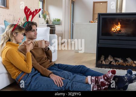 Couple tenant des tasses en céramique et portant des bandeaux en bois assis près de la cheminée dans le salon. Cosy, intimité, style de vie, scandine Banque D'Images